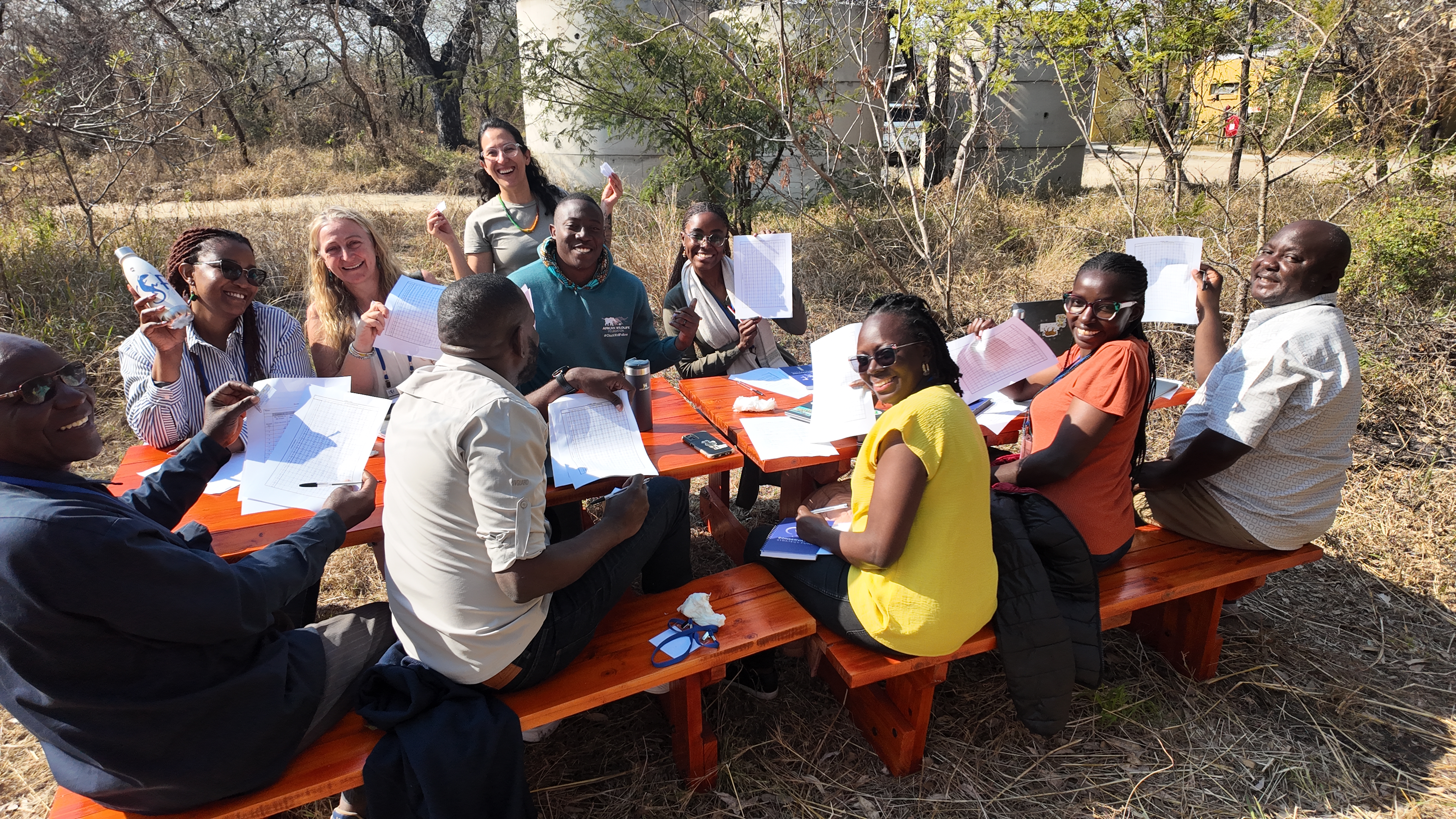 Participants smile and hold up their worksheets and game materials outside around two picnic benches.