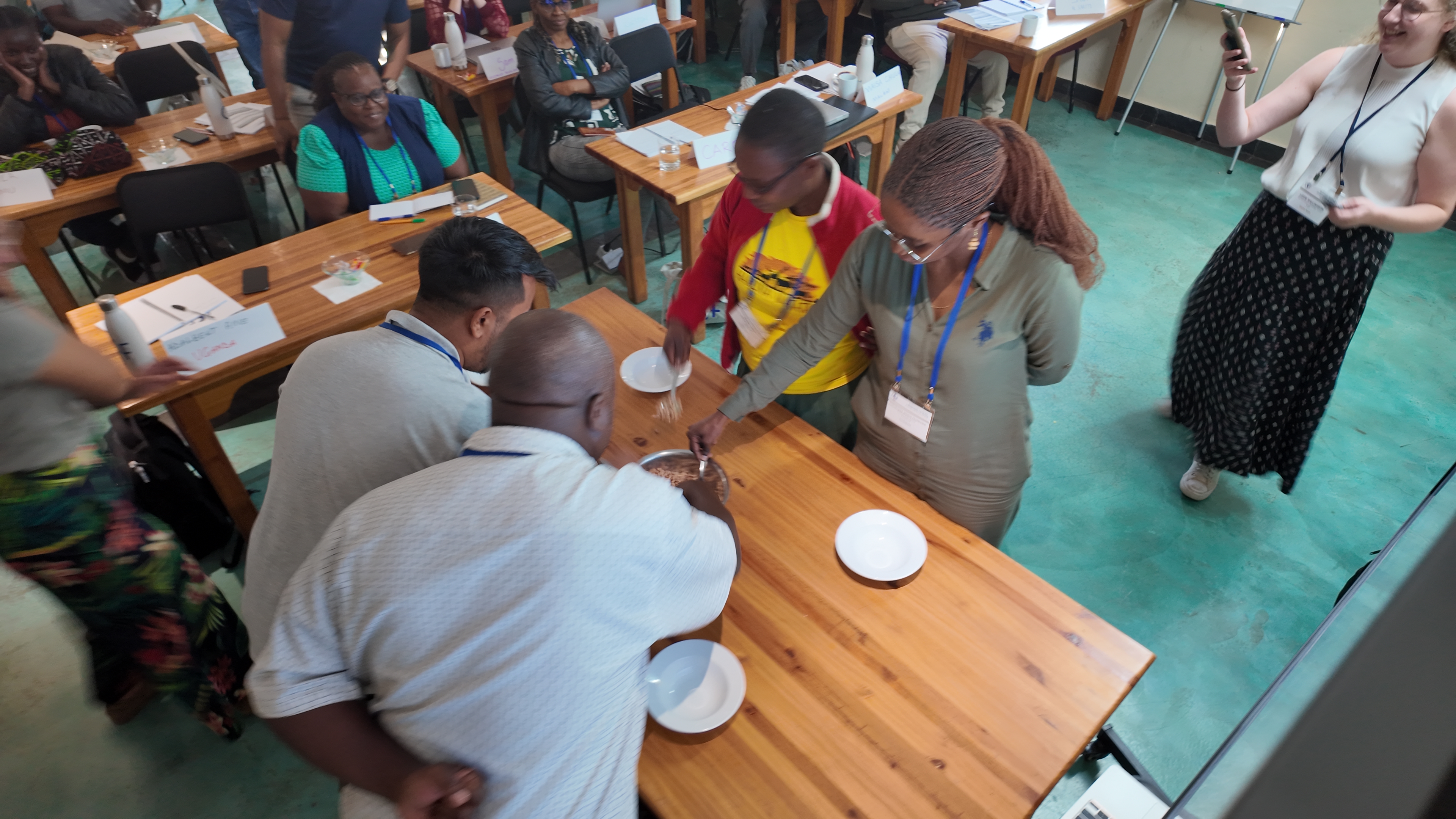 Four participant circle a table with spoons as they try to scoop beans out of a central metal bowl into their own, smaller bowls.